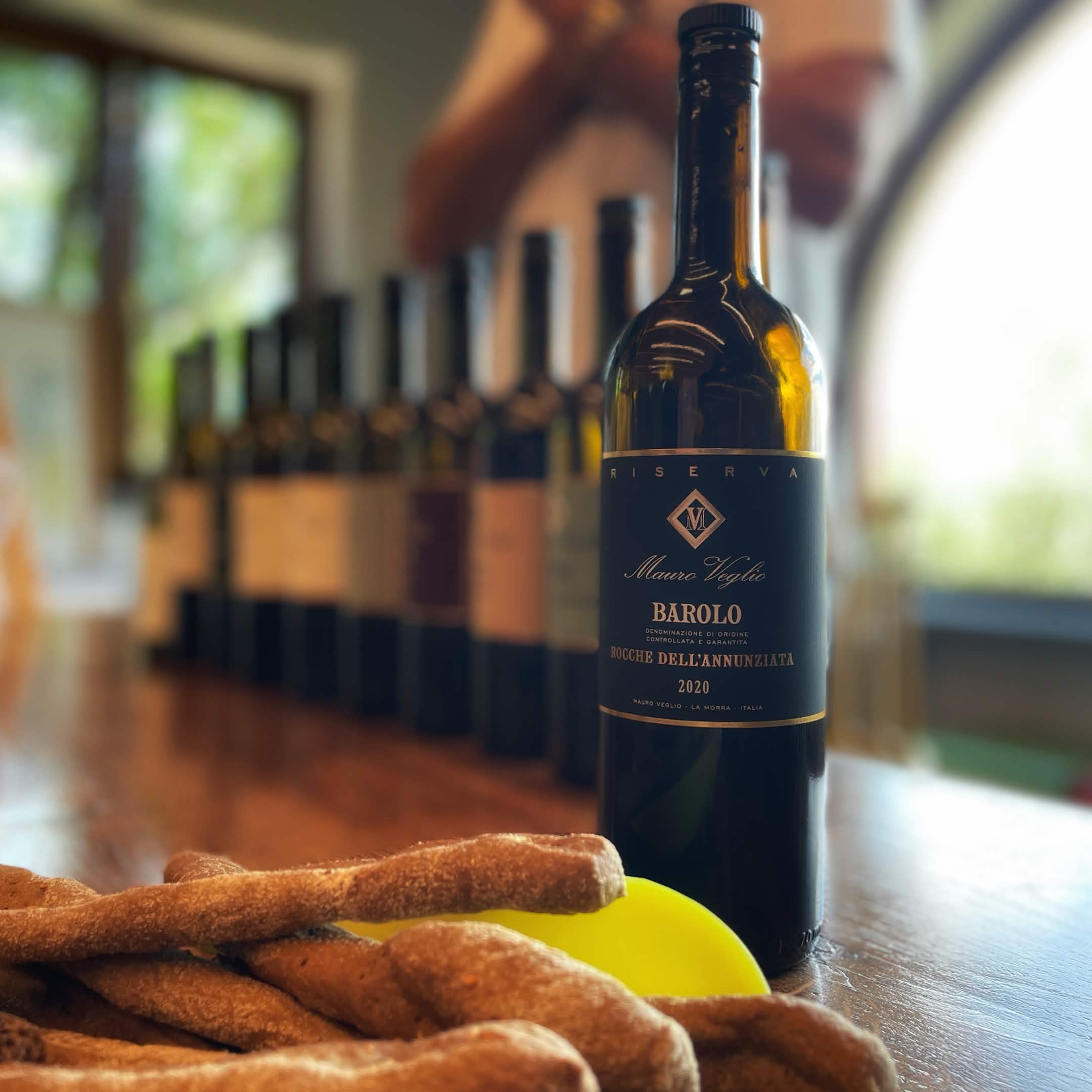 Fles Barolo-wijn op een houten tafel, met broodstengels en meerdere wijnflessen onscherp op de achtergrond